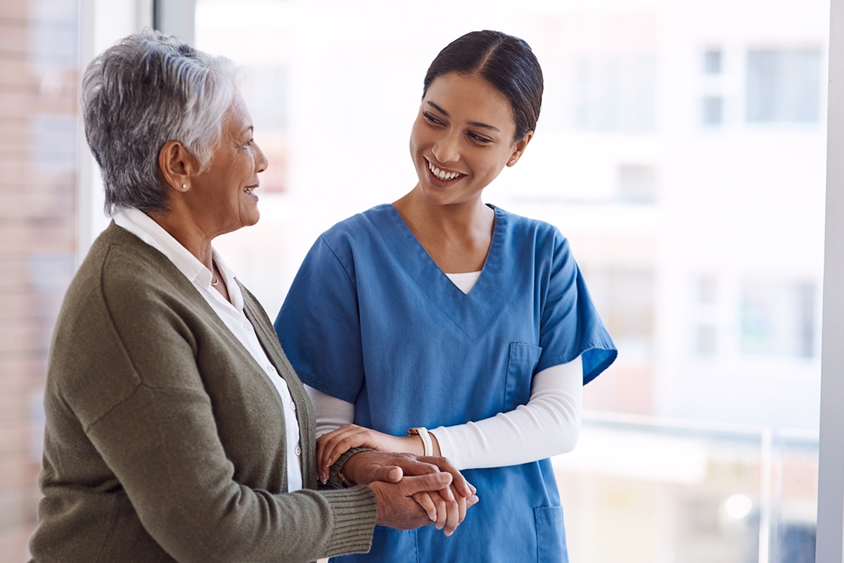 Support, caregiver with senior woman and holding hands for care indoors. Retirement, consulting and professional female nurse with elderly person smiling together for healthcare at nursing home